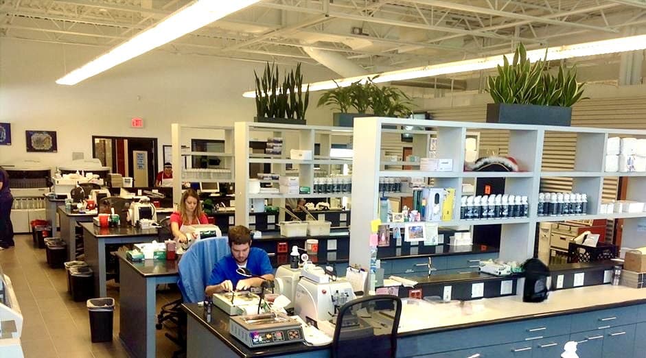 A wide angle view of the lab with rows of tables and shelves and two people working at two of the tables.