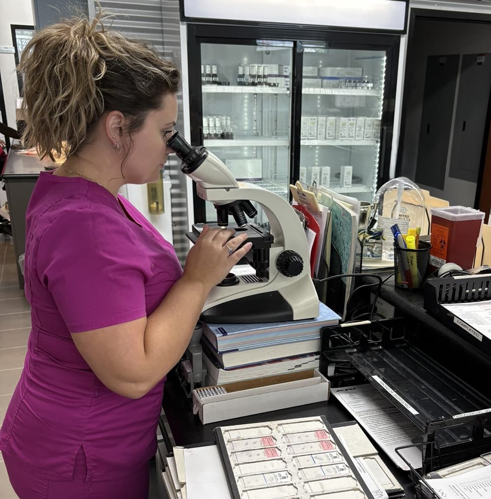 A woman with scrubs on standing at a table and looking through a microscope.