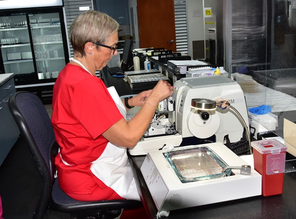 Woman with apron on sitting at a table with lab exam equipment on it. She is holding something in her hand.